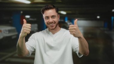 Arka planda bulanık arabalarla kapalı bir otoparkta dikilirken sakallı genç adam gülümsüyor ve başparmak kaldırıyor..