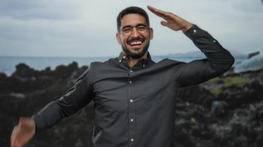 Sahilde gülümseyen, kayalık bir geçmişi olan, açık hava havası yayan, İspanyol cazibesini sergileyen, gözlük takan, plajın keyfini çıkaran genç bir adam..