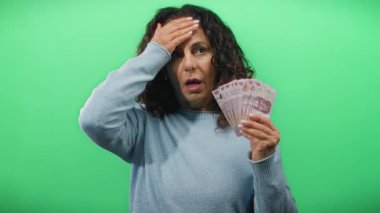 Orta yaşlı bir kadın, yeşil bir arka plan duvarına karşı Meksika pezosu tutuyor. Finansal kaygıları ve para yönetimi tasvir ediyor..