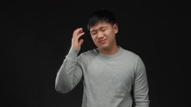 Gri kazaklı genç Çinli adam siyah bir arkaplan karşısında duruyor. Acı dolu bir ifadeyle kafasına dokunmaktan rahatsız olduğunu ifade ediyor..