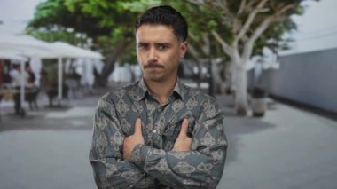 Şehrin caddesinde kendine güvenen genç bir adam çapraz kolları ve arka planda açık havada şehir ağaçlarıyla duruyor..