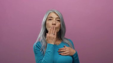 Mavi kazaklı yaşlı kadın pembe bir arka plana öpücük yolluyor jesti ve yüz ifadesiyle sıcaklığını ve sevgisini iletiyor..