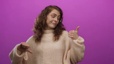 Gözlüklü ve süveterli bir kadın pembe arka planda parmaklarını öne ve arkaya gösteriyor. Canlı bir stüdyoda izole edilmiş neşeli bir ifadeyle karışık duygular ifade ediyor..