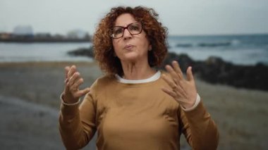 Woman gesturing with hands in city street wearing glasses, expressing with curly hair, hispanic middle-aged, outdoors by water, showing animated emotion and casual style.