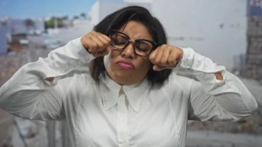 Gözlüklü genç bir kadın, bir inşaat alanında ağlama hareketi yapıyor, dışarıda duygularını ifade ediyor, Latin, artı beden, beyaz gömlek giyiyor..