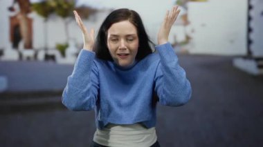 Esmer saçlı bir kadın sokak arka planında şaşkınlık ve tefekkür içinde, mavi bir süveter giyiyor, duyguları ve modayı somutlaştırıyor..