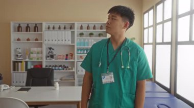 Klinikte kolları çapraz duran, önlük ve steteskop takan genç bir doktor yeterli ışık alan iyi donanımlı bir hastane ortamında kendine güvenini gösteriyor..