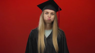 Mezuniyet kıyafetli genç bir kadın canlı kırmızı bir duvara karşı duruyor, neşeli bir duygu ifade ediyor, geleneksel akademik başarı üzerine neşeli bir bakış açısı sergiliyor..