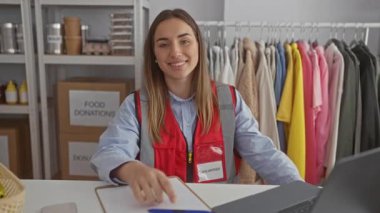 Bağış merkezinde masada oturan gönüllü bir kadın. Etrafı giysi ve yemek malzemeleriyle çevrili. Kapalı alanda kırmızı yelek giymiş kameraya gülümsüyor..
