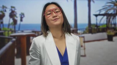 Gözlüklü ve beyaz ceketli genç Çinli kadın, arka planda palmiye ağaçları ve okyanus manzaralı bir şehir restoranının terasında güvenle duruyor..