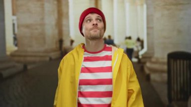 Sarı yağmurluk ve kırmızı bereli genç adam şehir sokaklarında neşeli bir şekilde el kol hareketi yapıyor. Açık hava kentsel ortamının ortasındaki şehir canlılığını ve gençlik enerjisini somutlaştırıyor..