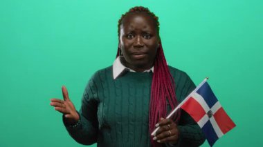 Yeşil arka planda Dominik bayrağını tutan, duygularını el hareketleriyle ifade eden, renkli süveter ve örgü takan, Dominik mirasını ve kültürel gururu sembolize eden bir kadın..