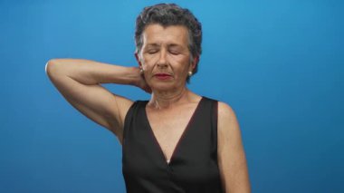 Mavi arka planda siyah giysili, gri saçlı yaşlı kadın, boyun ağrısını ifade ediyor, gözleri kapalı, rahat duruşu var, rahatsız ama sakin duruşu var..
