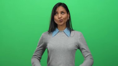 İzole edilmiş yeşil bir duvarın önünde, mavi yakalı gri bir kazak giyen eğlenceli ve genç bir kişilik sergileyen bir kadın..