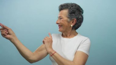 Gri saçlı yaşlı kadın mutlu bir şekilde mavi arka planı işaret ediyor, neşe ve kendine güveni ifade ediyor..