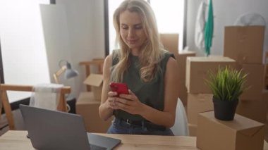 Yeni evin oturma odasında dizüstü bilgisayarı ve akıllı telefonuyla gülümseyen bir kadın yeni taşındığını gösteren kutularla çevrili..