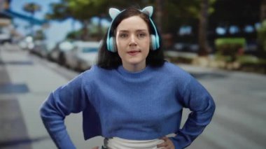 Kulaklık takan ve mavi kazak giyen bir kadın güneşli bir sokak sahnesinde arka planda ağaçlar ve arabalar olan ciddi bir ifadeyle el kol hareketi yapıyor..