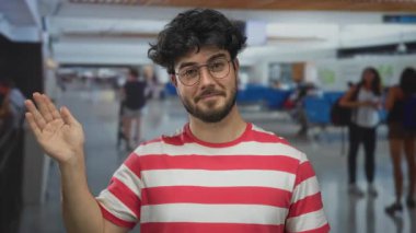 Sakallı ve gözlüklü genç bir adam, çizgili bir gömlek giyiyor, arka planda bulanık gezginlerle dolu kapalı bir havaalanında kameraya el sallıyor..
