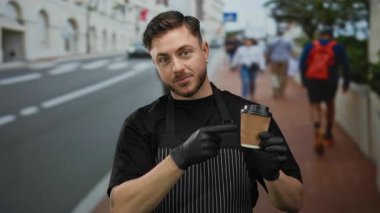 Önlüklü genç adam, şehir caddesinde beklerken kahve fincanına işaret ediyor. Açık havada eldivenli garson rolünün altını çiziyor..