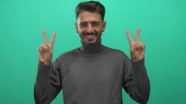 Genç İspanyol adam iki eliyle barış işareti gösteriyor ve stüdyoda yeşil duvara karşı gülen yüz; iyimserlik..
