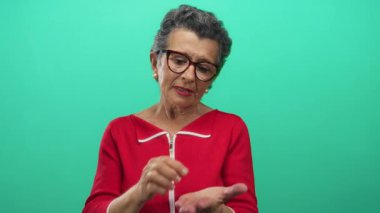 Kırmızı tişörtlü, gri saçlı yaşlı bir kadın yeşil arka planda para için el kol hareketi yapıyor. Zorlayıcı bir görsel tasvirle maddi gereksinim ve bağımsızlık taşıyor..