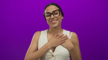 İzole edilmiş mor arka planda gülümseyen bir kadın mutluluğu ve pozitifliği ifade eder, gözlük takar ve beyaz bir bluz giyer, sıcaklığı ve arkadaşlığı kendinden emin bir şekilde ifade eder..