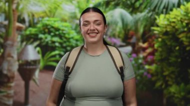 Güneşli bir parkta gülümseyen ve eğlenen bir kadın canlı yeşil bitkilerle çevrili, doğanın güzelliğini kucaklarken mutluluk ve sükunet saçıyor.