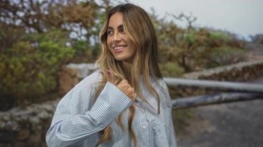Sarışın, çizgili gömlek giyen bir kadın ormanda baş parmak kaldırıyor. Onaylama iyimserliği, güven sevinci..