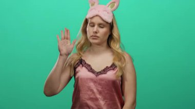 Pembe gecelikli, yeşil arka planda uyku maskesi takmış bir kadın el kaldırıyor. Kapalı gözler ve sakin bir ifadeyle..