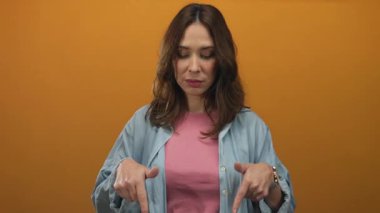 İzole edilmiş turuncu arka plana bakan bir kadın genç İspanyol kadını işaret ediyor. Ciddi ifade. Sıradan giyinen kot ceket kolları el ele..