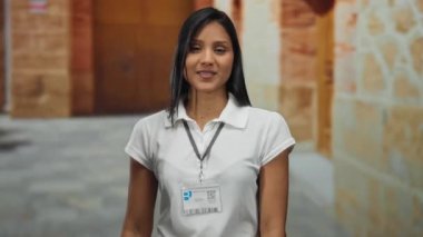 Beyaz tişörtlü, kimliği olan genç bir İspanyol kadın üniversite binasının arka planını gösterirken gülümsüyor..