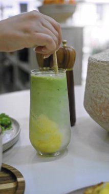 Kafeterya masasında el çırpması yeşil smoothie. Tuz makinesinin yanında ahşap sopa ve taze, sağlıklı bir içecek seti sergiliyor.