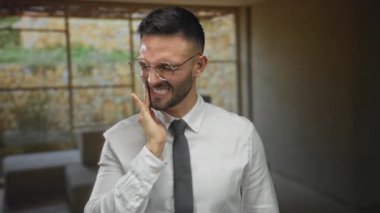 Kırsal bir Akdeniz evinde gözlüklü genç bir adam diş ağrısı çekiyor gibi görünüyor, resmi bir gömlek ve kravat takıp arka planda bulanıklık yaratıyor..