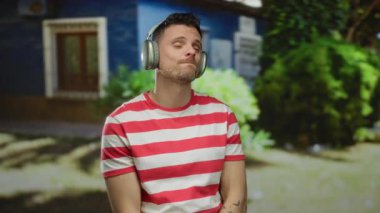 Kulaklık takan genç bir adam şehir ortamında müzik dinlemekten hoşlanır, çizgili bir gömlek giyer, yeşillik ve binalarla çevrilidir..