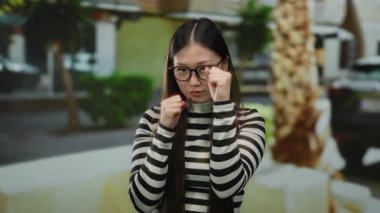 Gözlüklü, neşeli bir boks pozu veren, arka planı bulanık, çizgili gömlekli bir kadın..