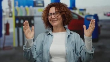 Orta yaşlı İspanyol kadın, arka planda arabalarla benzin istasyonunda el kol hareketleri yapıyor, dış mekanda ifade ve etkileşim sergiliyor..