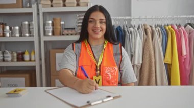 Bağış ve kıyafetlerle çevrili bir hayır kurumunda yazan bir kadın, güvenlik yeleği ve gönüllü kimlik takıyor, gülümsüyor ve toplumsal işleriyle meşgul..