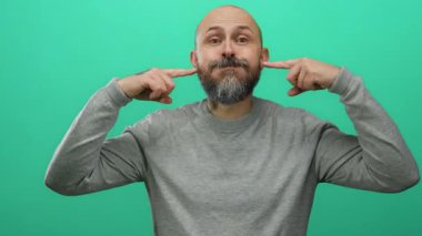 Sakallı kel adam izole edilmiş yeşil bir duvarın önünde neşeli tavırlarını ve günlük tarzını vurgulayarak neşeli ifadeler sergiliyor..