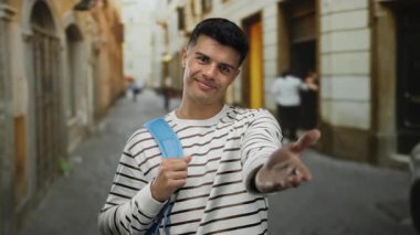 Çizgili gömleği ve mavi sırt çantasıyla yakışıklı genç bir adam şehrin arka planında dostluk ve gündelik tarzını gösteren bir el uzatıyor..