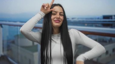 Kaybeden kadın yolcu teknesinde arka planda okyanusla işaret veriyor, beyaz gömlek giyiyor, güneşli bir günde açık deniz manzarasını seyrediyor, neşeli ve genç bir enerji saçıyor..