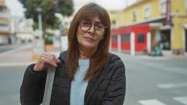 Şehrin sokak ortamında güvenle gülümseyen İspanyol asıllı son sınıf öğrencisi kadın, şehir arka planında, açık havada yakalanmış, açık renk süveter, gözlük ve gündelik ceket, somut modern yaşam tarzı..