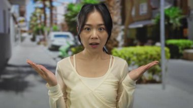 Genç Çinli kadın güneşli bir caddede dikiliyor. Arka planda palmiye ağaçları ve şehir sokaklarında park edilmiş arabalarla sürpriz yapıyor..