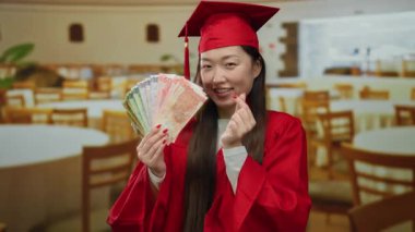 Mezuniyet elbisesi giymiş, elinde Filipin pesoları olan bir kadın bir restoranda gülümseyerek başarı ve eğitim sergiliyor..