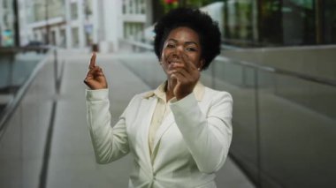 Beyaz takım elbiseli kadın, arka planda binalar olan bir şehir caddesinde gülümsüyor ve jestler yapıyor..