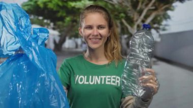 Kadın çantası taşıyan gönüllü, çevrede gülümserken plastik şişe ve mavi çöp temizliyor..