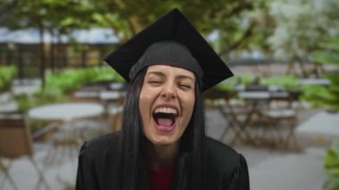 Mezuniyet şapkası ve cübbesi içinde ağaçlarla çevrili bir teras kafesinde gülümseyen bir kadın, başarısını neşeli bir ifadeyle kutluyor..