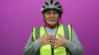 Kask ve yansıtıcı yelek giymiş, aktif yaşam tarzı ve pozitifliği somutlaştıran, izole edilmiş pembe bir arka plana karşı kollarını açmış, güvenlik donanımlı İspanyol kıdemli kadın..