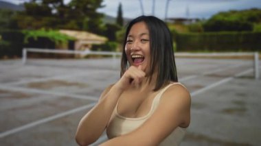 Yeşil ve manzaralı arka planla çevrili açık tenis kortunda güvenle gülümseyen genç bir kadın eğlenceli bir atmosfer olduğunu gösteriyor..