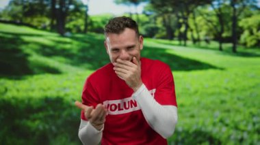 Gönüllü gömlekli genç adam parkta yemyeşil ağaçlar ve çimlerle çevrili güneşli bir günün tadını çıkarırken gülüyor.