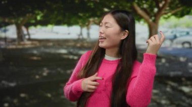 Pembe kazaklı bir kadın ağaçlarla dolu bir parkta neşeli bir şekilde gülüyor ve güneşli bir günün tadını çıkarıyor..